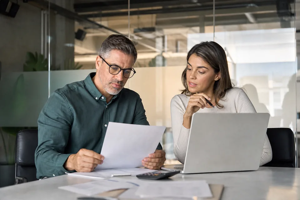Two data leads comparing a monitoring vendor quote on paper and a laptop in a meeting room